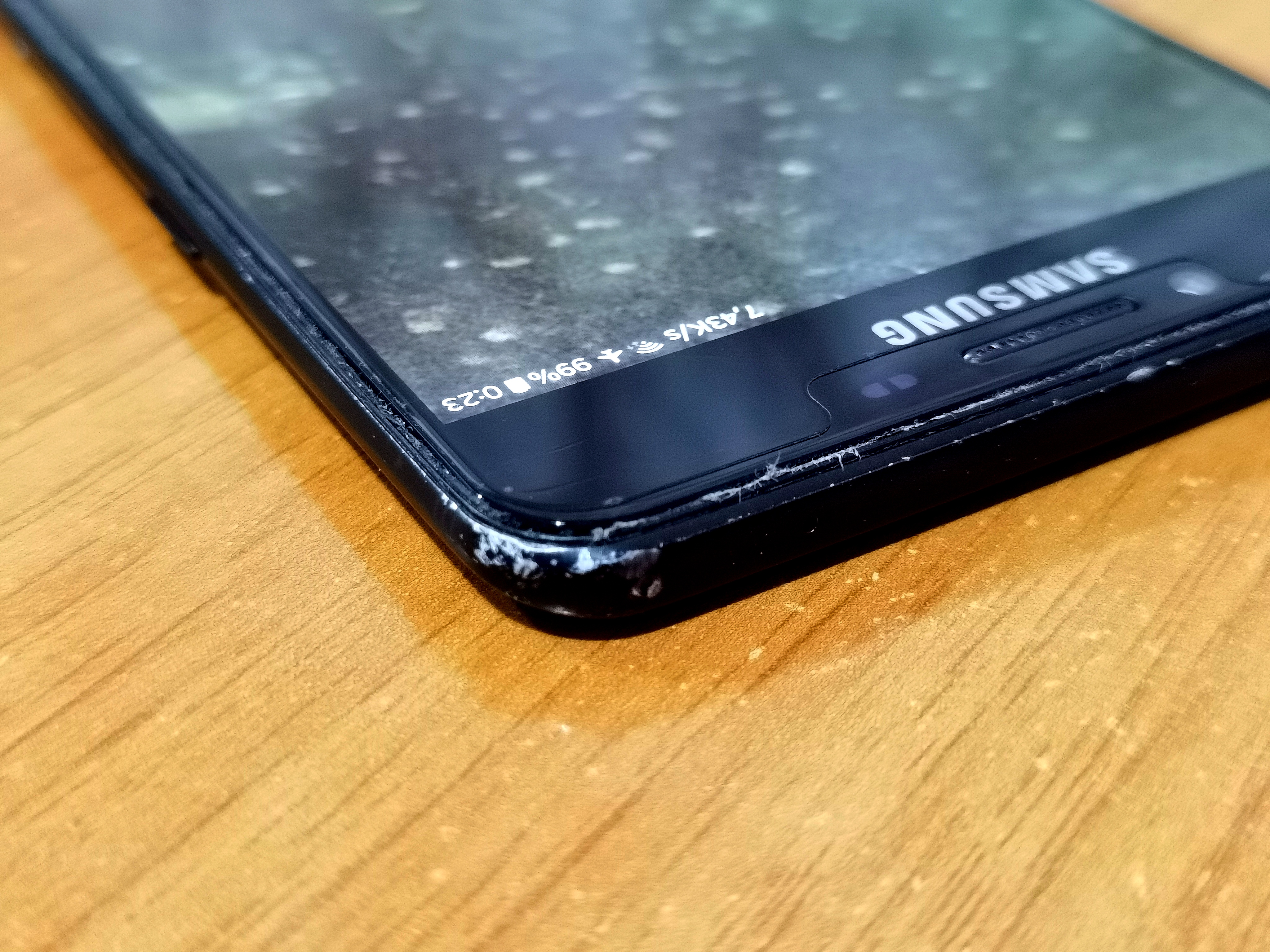 a closeup of a corner of the phone showcasing the frame and screen and the glue inbetween just slightly coming off, the screen is on the home screen with a black garden wallpaper, the top bar is visible and you can see the phone has 99% of battery left and the clock showing 0:23